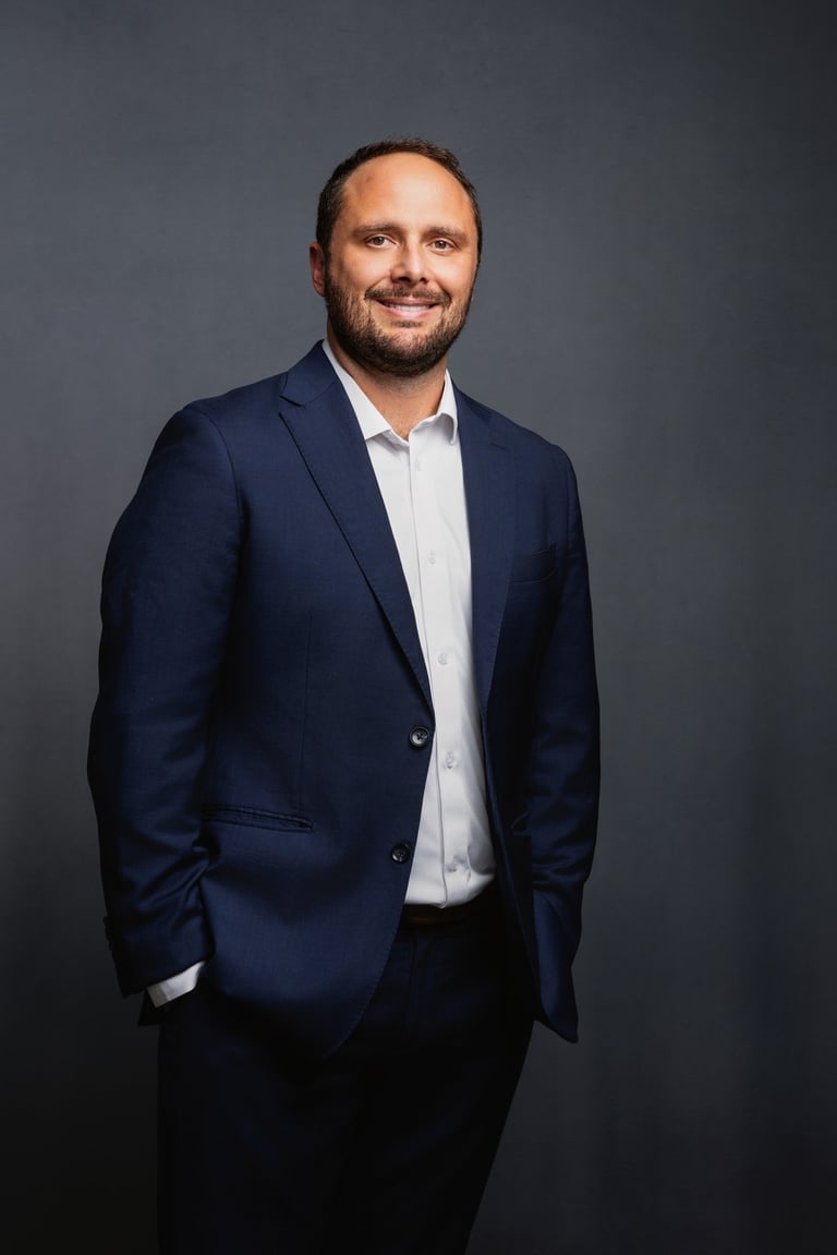 Professional headshot of a bearded man in a dark navy blazer and white shirt, smiling against a gray background.